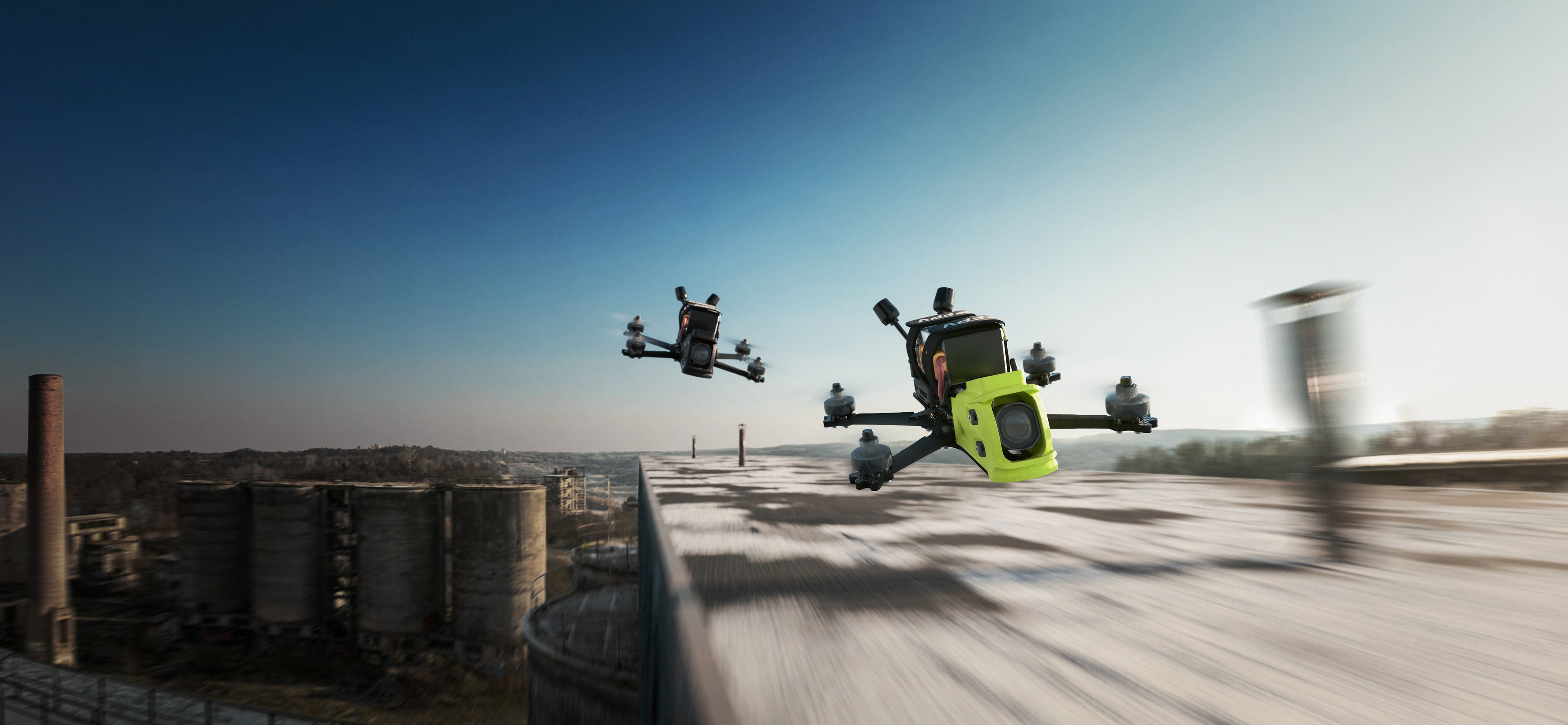 Action shot of two It's FPV ERA Mini freestyle fpv drones flying fast over an abandoned industrial roof. The lead drone features a  bright lime camera cage, followed by a black model against a blue sky.